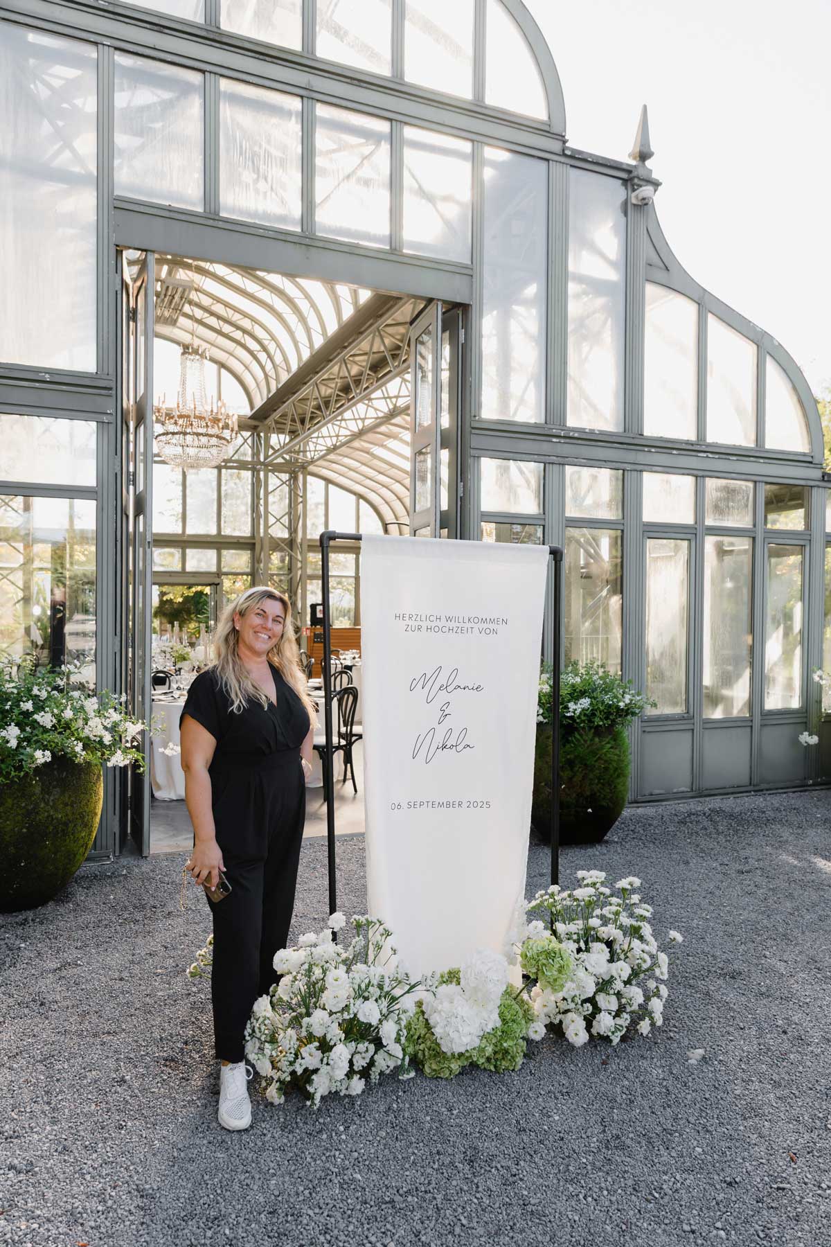 Vor einem hellen Glashaus steht eine Frau in Schwarz neben einem Willkommensschild, umgeben von weißen und grünen Blumen.