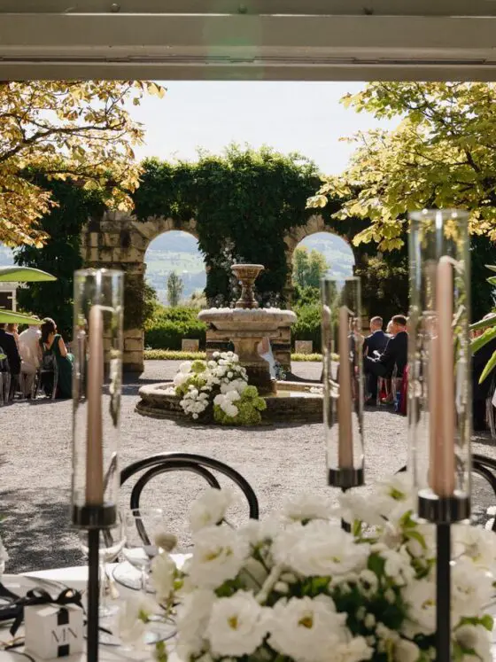 Ein sonniger Garten mit Steinbrunnen, weißen Blumenarrangements und begrünten Bögen im Hintergrund.