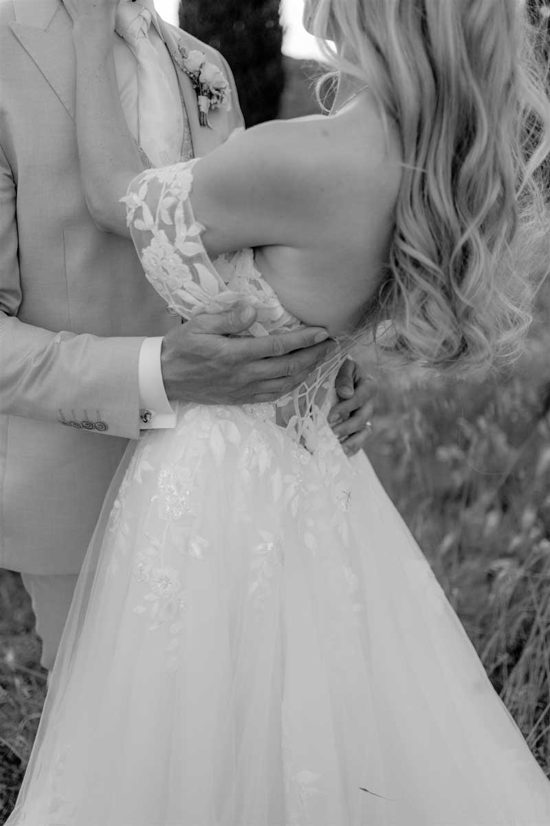 Eine Braut mit besticktem Kleid lehnt sich an den Bräutigam, der sie im Freien behutsam an der Taille hält.