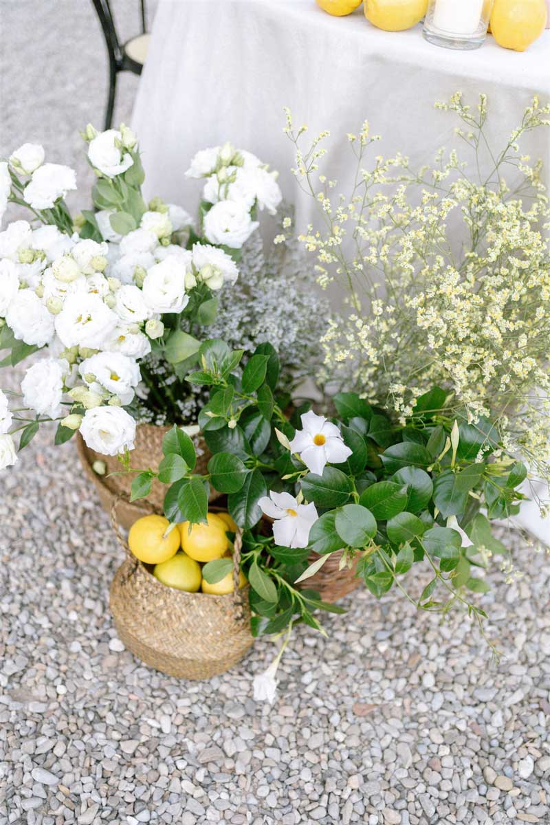Weiße Blüten, grünes Blattwerk und gelbe Zitronen stehen in geflochtenen Körben auf hellem Kies vor einem hellen Tuch.