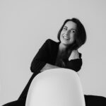Mit einem offenen Lächeln sitzt Alica Wagner in schwarzer Kleidung vor einem hellen Hintergrund auf einem weißen Stuhl.