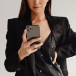 Eine Frau im schwarzen Blazer hält ein Smartphone vor dem Oberkörper, vor hellem, ruhigem Hintergrund.