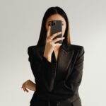Vor hellem Hintergrund macht eine Frau im schwarzen Blazer ein Spiegel-Selfie und verdeckt ihr Gesicht mit dem Smartphone.