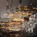 Warme Lichtreflexe treffen auf zwei gefüllte Cocktailgläser, Spiegelkugeln und glitzernde Deko auf einem dunklen Tisch.