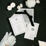 Weiße Hochzeitspapeterie mit schwarzen Schleifen, goldenen Akzenten, Ringen, Handschuhen und weißen Blüten auf dunklem Grund.