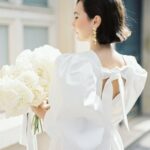 Eine Braut mit kurzem schwarzem Haar trägt ein weißes Kleid mit Puffärmeln und einen großen Strauß weißer Blüten am Fenster.