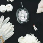 Auf schwarzem Grund liegen ein Diptyque-Flakon L’Eau Papier, weiße Handschuhe, Schmuckschachteln und eine große weiße Blüte.