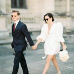 Ein Brautpaar geht Hand in Hand über helles Pflaster; sie trägt ein kurzes weißes Kleid, er einen dunklen Anzug mit Sonnenbrille.