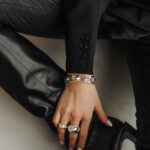 Zu sehen ist eine Hand mit silbernen Ringen und Armbändern zu einem schwarzen Outfit aus Blazer, Lederhose und Schuhen.
