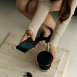 Alena Scheffler hockt neben einem hellen Steintisch, hält Smartphone und Becher und trägt einen beigefarbenen Strickpullover.