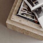 Auf einer hellen Travertinablage liegen mehrere aufgeschlagene Magazine mit Schwarzweiß- und Farbfotos in warmem Licht.