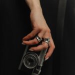 Vor dunklem Hintergrund hält eine Hand eine silberne Kamera, die durch große Ringe und helle Nägel auffällt.