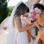 Eine Braut im weißen Kleid mit Schleier hält einen pastellfarbenen Blumenstrauß, daneben steht eine Begleiterin in Orange.