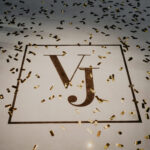 Goldenes Konfetti liegt auf hellem Boden, in der Mitte steht ein eingerahmtes Monogramm mit den Buchstaben VJ.