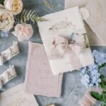 Feine Hochzeitskarten, ein helles Kuvert, Seidenband und Blumen liegen auf graublauem Stoff in Pastellfarben.