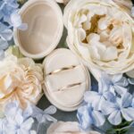 Zwei Eheringe liegen in einer cremefarbenen Ringbox, umgeben von hellen Blumen in sanften Pastelltönen.