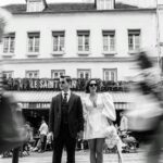 Ein Brautpaar steht in Schwarzweiß vor dem Café Le Saint, während unscharf vorbeigehende Passanten die Straßenszene beleben.