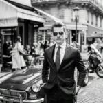 Ein Mann im dunklen Anzug und mit Sonnenbrille steht vor einem Oldtimer auf einer belebten Altstadtstraße.