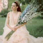 Eine Braut in zartem Tüllkleid hält ein langes Bouquet aus weißen und blauen Blüten vor einer weichen grünen Kulisse.