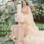 Brautpaar in zarten Pastelltönen steht vor einer mehrstöckigen Hochzeitstorte, umgeben von üppigen Blumen und Gartentor.