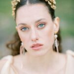 Nahes Porträt einer jungen Frau mit blauem Blick, goldenem Haarschmuck und hellem, weichem Hintergrund.