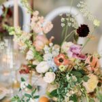 Pastellige Blumenarrangements mit Kerzen, zarten Wildblüten und Glasvasen schmücken eine elegant gedeckte Hochzeitstafel.