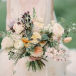 Ein pastellfarbener Brautstrauß mit Rosen, Tulpen und zarten Wildblüten vor grünem Hintergrund und hellem Stoff.