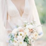 Eine Braut in einem luftigen Kleid mit tiefem Ausschnitt hält einen pastellfarbenen Strauß aus weißen, roséfarbenen und blauen Blüten.