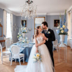 Ein Brautpaar steht in einem hellblauen Salon mit Kristalllüster, weißem Mobiliar und blauen Blumenarrangements vor einer Tafel.