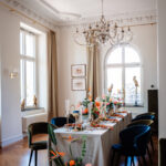 In einem hellen Salon steht ein lang gedeckter Tisch mit Blumen, Kerzen und Veloursstühlen unter einem großen Kristalllüster.