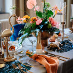 Ein festlich gedeckter Tisch mit hohem Blumenarrangement in Orange, Pink und Grün, Goldbesteck und einer orangenen Serviette.