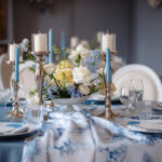 Ein festlich gedeckter Tisch zeigt ein blau-weißes Arrangement mit Blumen, Kerzen in Glaszylindern und feinem Geschirr.