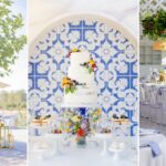 Vor einer blau-weißen Musterwand stehen eine mehrstöckige Torte, florale Arrangements und ein festlich gedeckter Tisch.