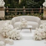 Weiße Blumen umrahmen ein helles Sofa mit rundem Tisch vor einer goldenen Gartenbalustrade im Freien.
