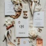 Helle Hochzeitsdetails liegen als ruhiges Flatlay auf einem beigen Untergrund, mit Karten, Schuhen, Blüten und Schmuck.