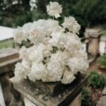 Weiße Blüten in einer üppigen Arrangementschale stehen auf einem verwitterten steinernen Sockel vor grünem Gartenhintergrund.