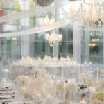 Lange Festtafel mit weißen Blumen, Kerzen und silbernen Dekoakzenten unter Glaspendeln im hellen Saal.