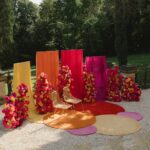 Bunte Blumenarrangements, farbige Stoffbahnen und goldene Stühle stehen in einer sonnigen Gartenkulisse.