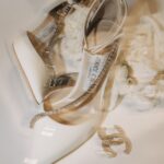 Weiße Brautschuhe mit spitzer Kappe und Kristallriemen liegen vor hellen Blüten, dazu schimmert ein goldener Innenbereich.
