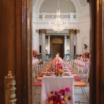 Durch eine geöffnete Holztür fällt der Blick in einen hellen Festsaal mit langer Tafel, Kronleuchter und pinken Blumen.