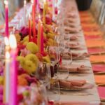 Eine lange festlich gedeckte Tafel mit bunten Kerzen, üppigen Blumen und vielen Gläsern in warmem Licht.