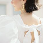 Eine Frau mit kurzem dunklem Haar trägt ein weißes Kleid mit Schleife am Rücken und goldene Blütenohrringe im weichen Licht.
