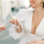 Eine lächelnde Braut im weißen Kleid hält ein Champagnerglas, am linken Rand ist eine weitere Hand mit Glas angeschnitten.