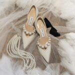 Cremefarbene Jimmy-Choo-Pumps mit Perlendetail liegen neben einer mehrreihigen Perlenkette auf hellem Tüll.